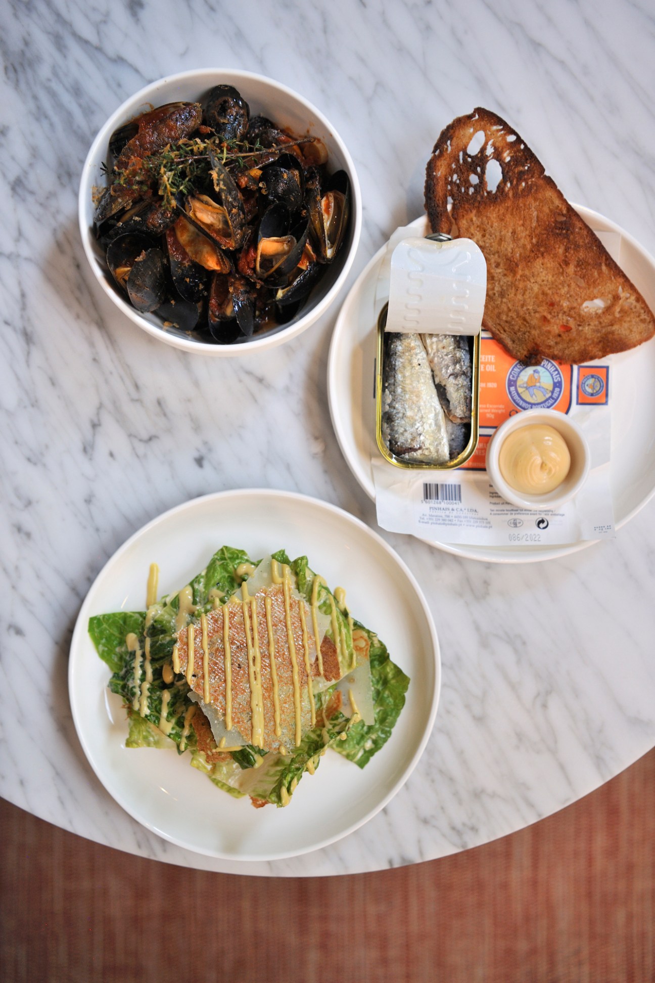 Overhead view of plated dishes including mussels, Caesar salad, canned fish, bread and sauce on marble surface.