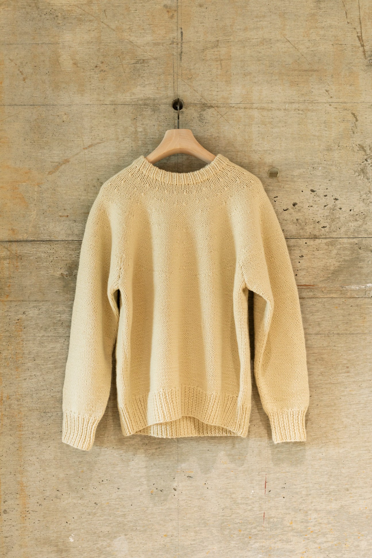 Cream-colored knit sweater hanging on wooden hanger against textured concrete wall.