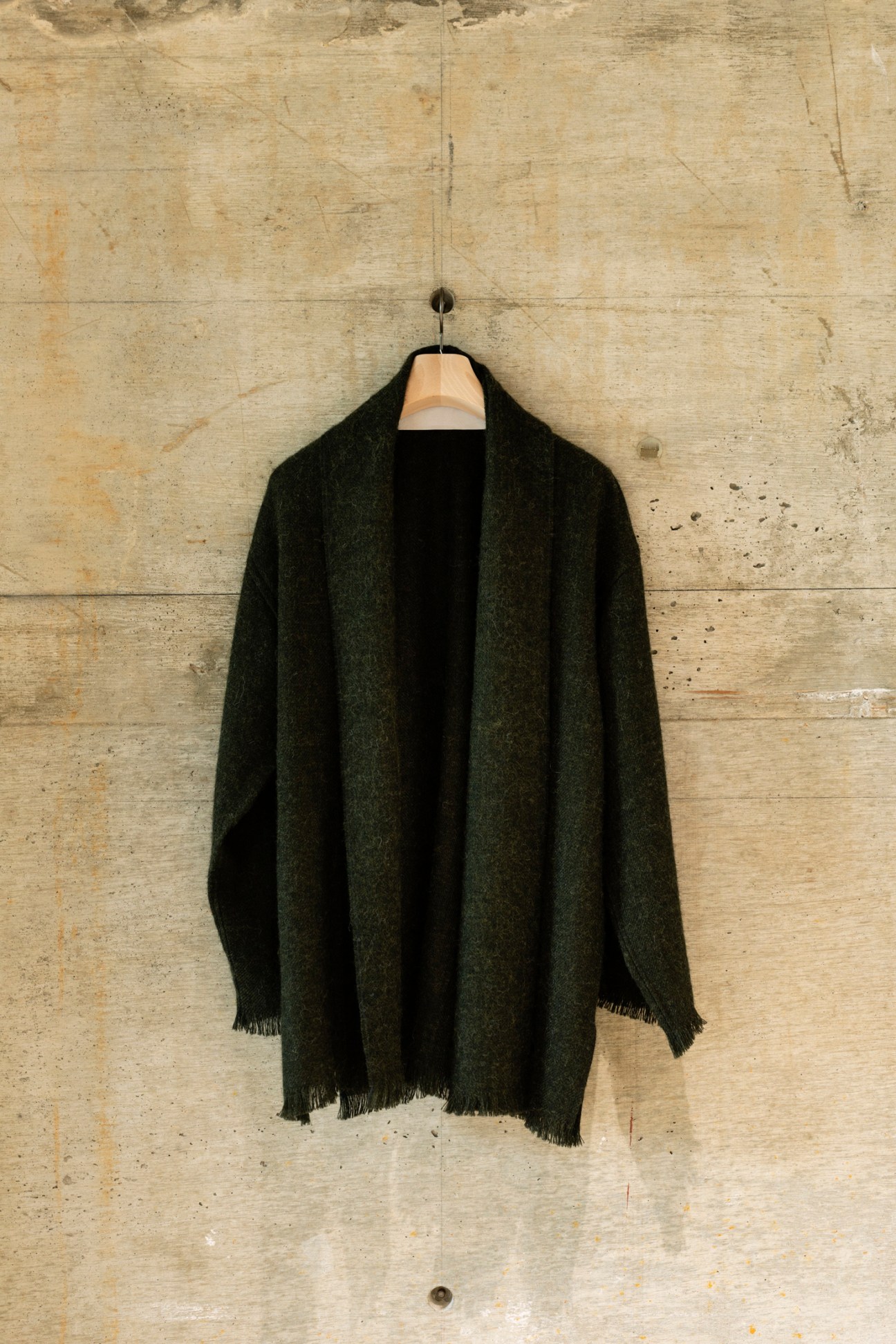 Dark charcoal cashmere sweater with frayed edges hanging on wooden hanger against concrete wall.
