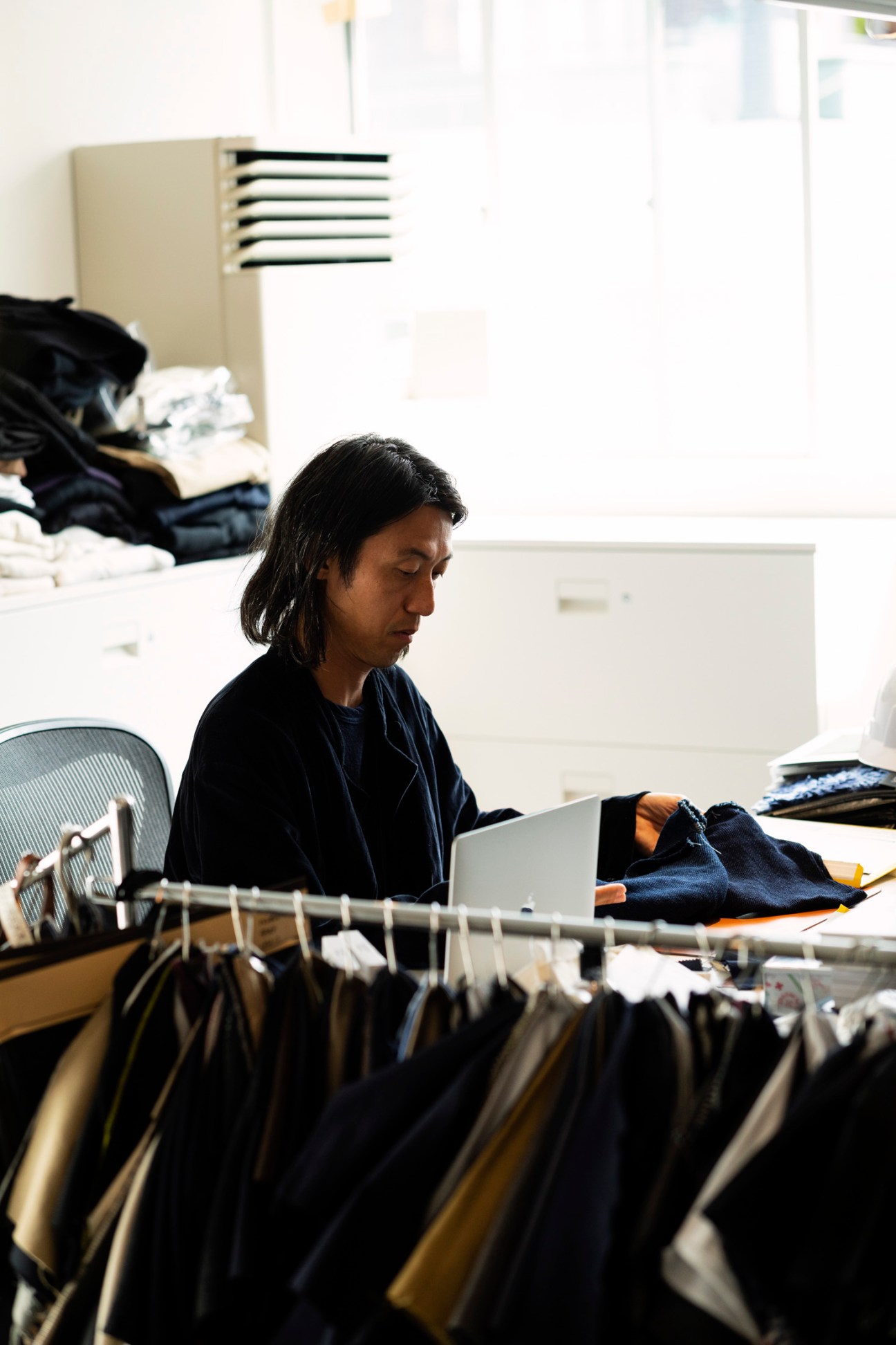 Designer Keijiro Komori sits at a desk working on a laptop, surrounded by clothing samples and fabrics in his studio.