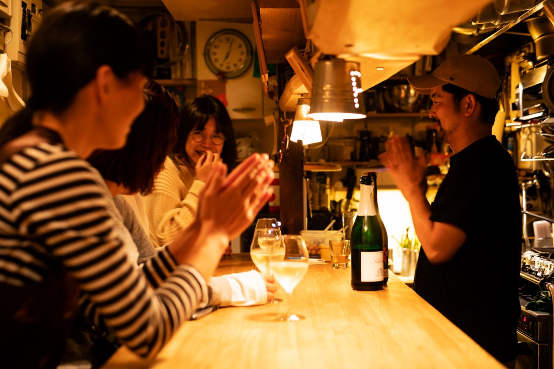 Customers applaud at a cozy Japanese bar counter with champagne bottle and warm lighting