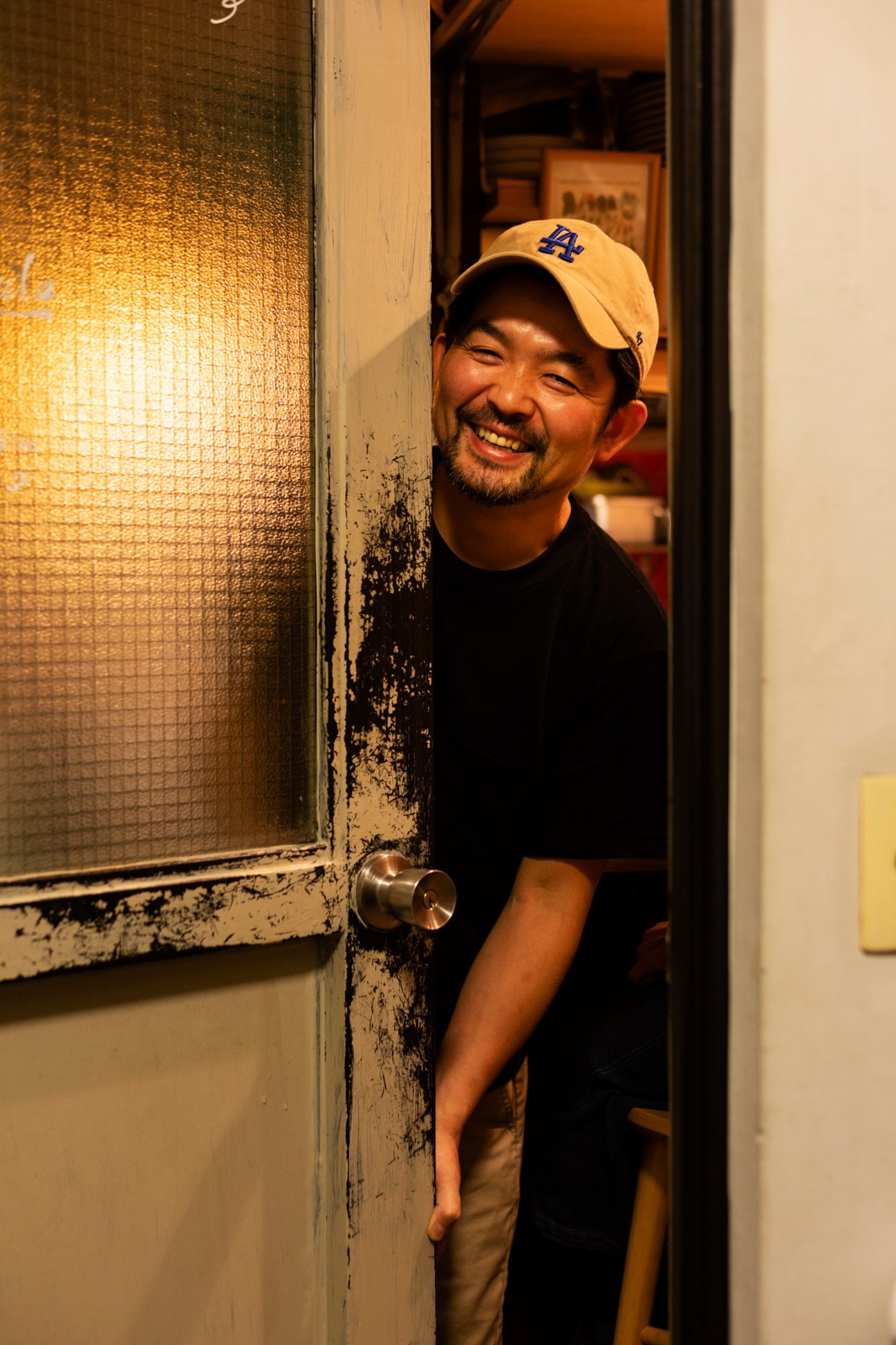 Smiling chef in LA cap and black shirt standing in doorway of restaurant kitchen
