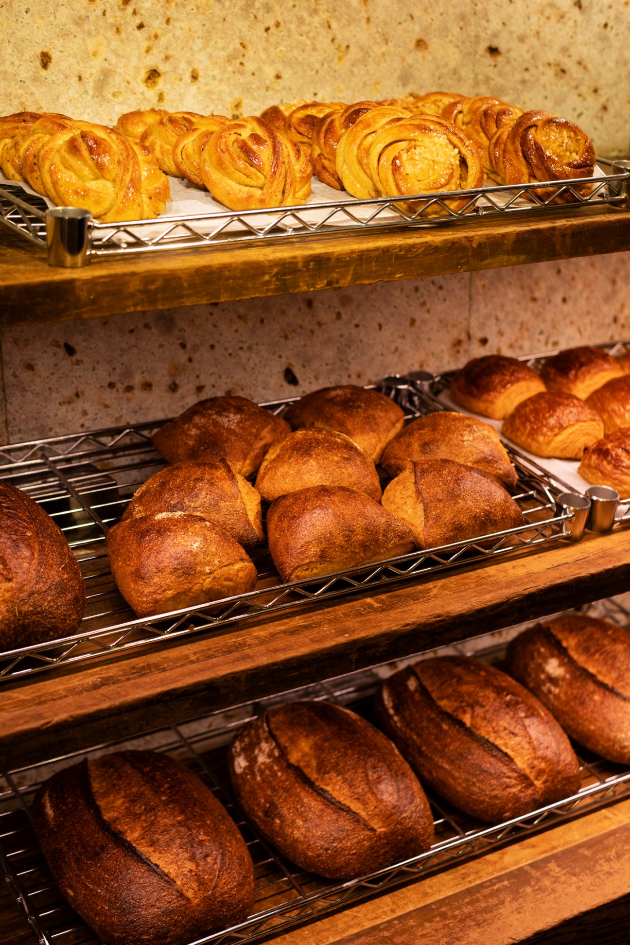 Freshly baked artisanal breads displayed on wire cooling racks in a bakery, showing various shapes and golden-brown crusts