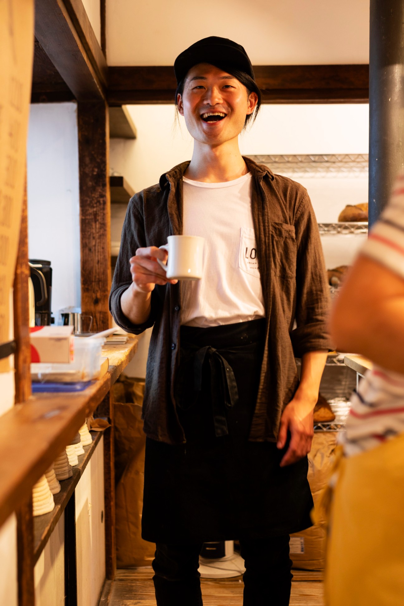 Smiling baker Tsukasa Miyawaki holds a white coffee cup in a rustic bakery kitchen with wooden beams and shelving