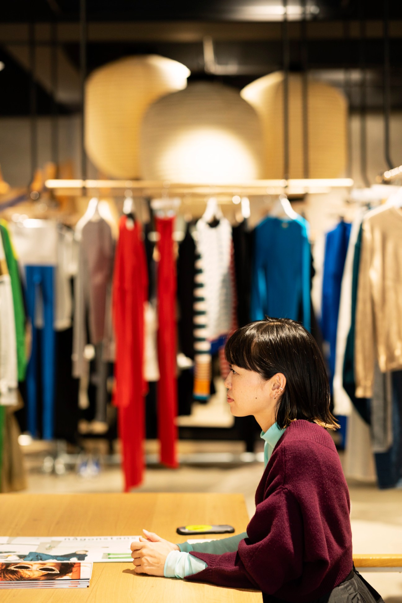 Creative director Mizuki Ueda sitting at a table in front of clothing racks displaying colorful garments.