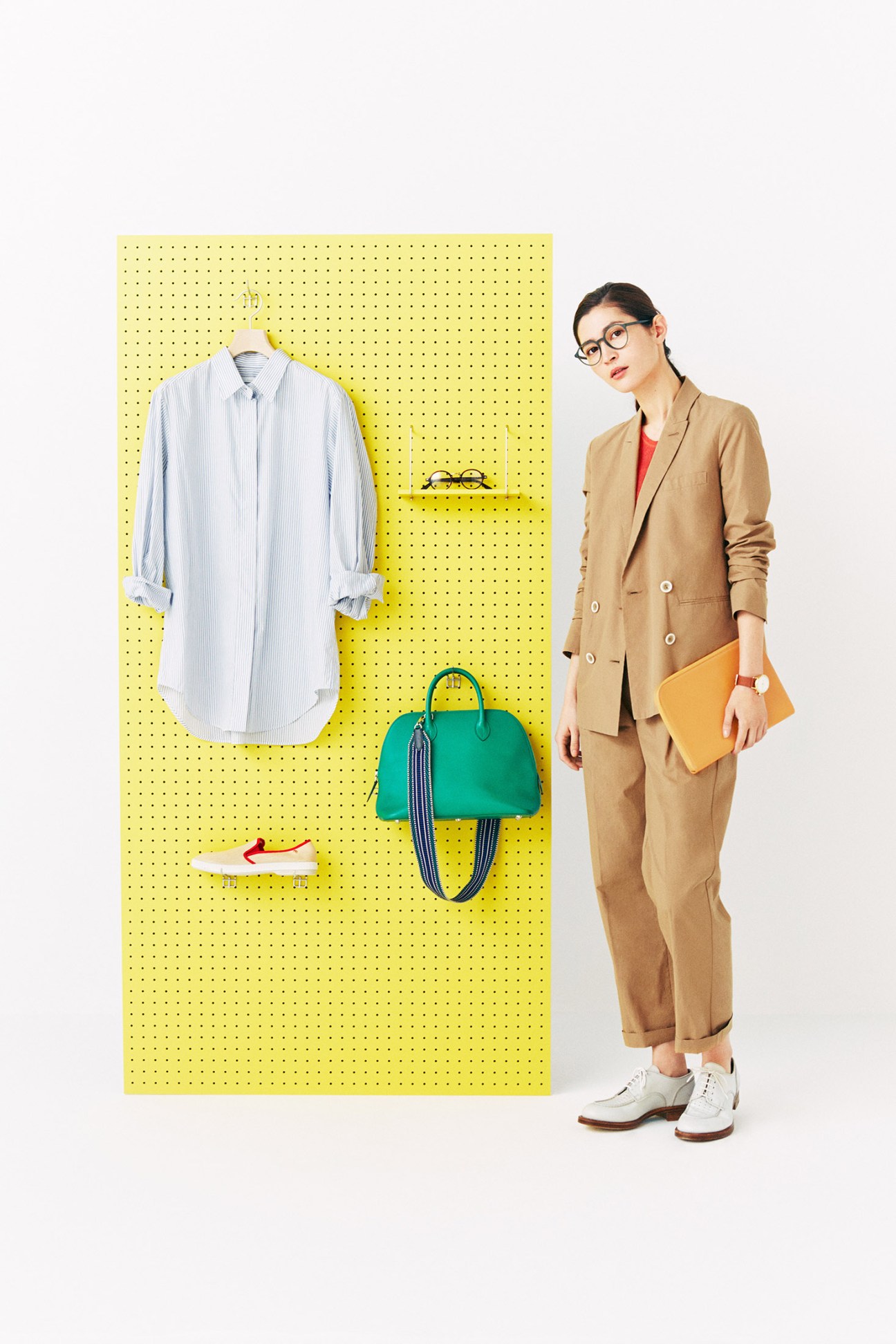 Woman in camel blazer and trousers stands beside yellow pegboard displaying spring wardrobe items.