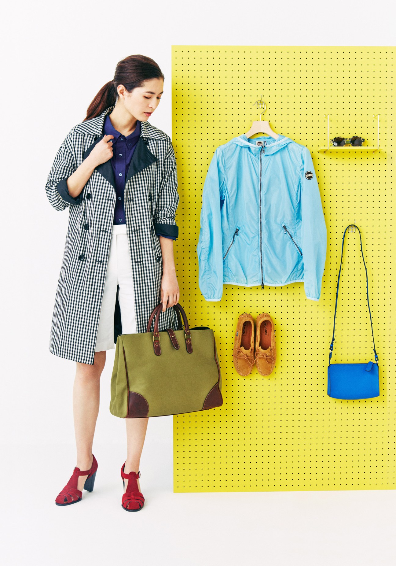 Woman wearing Herno coat and Ettinger bag poses next to yellow pegboard displaying spring clothing and accessories.