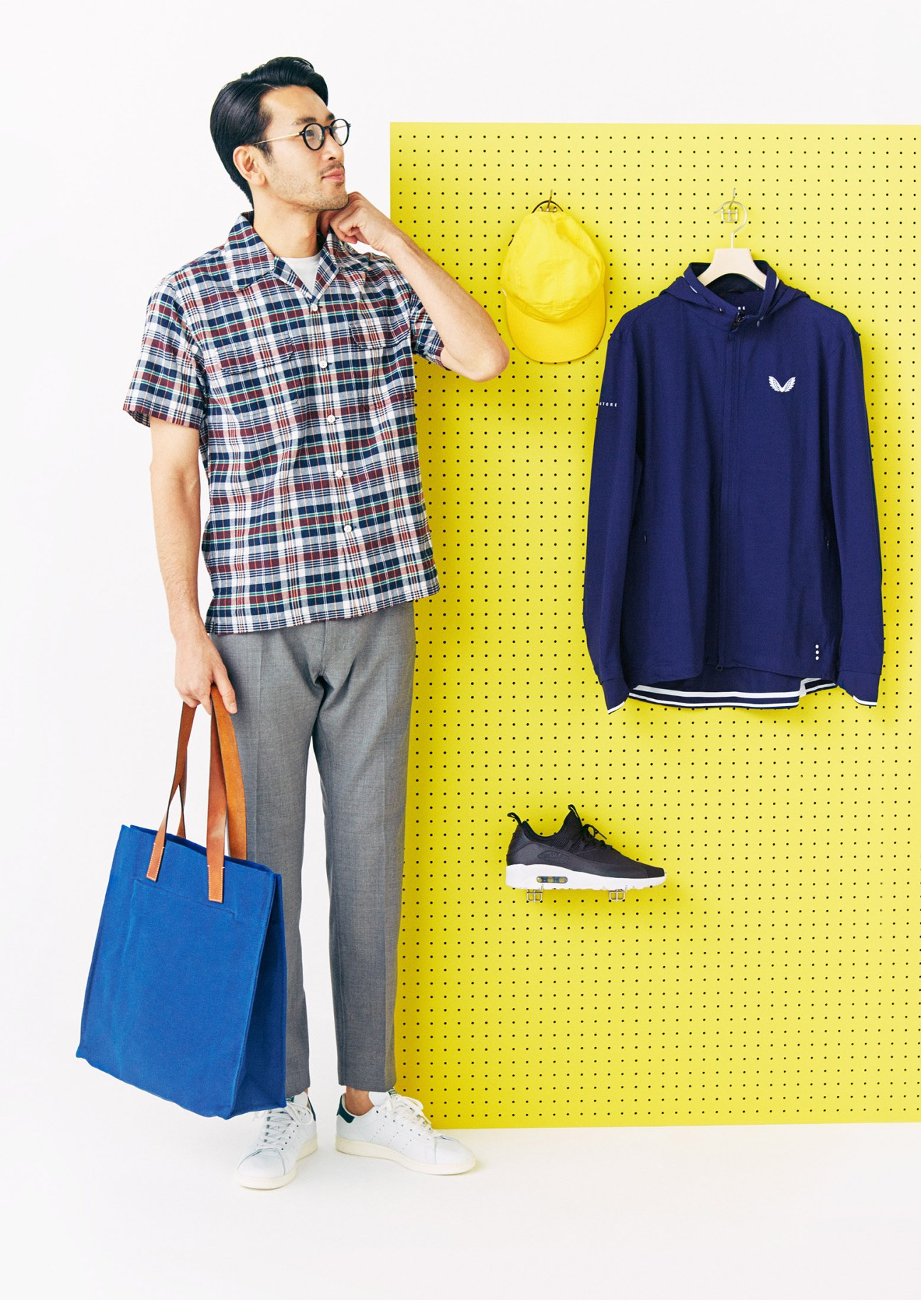 Male model wearing plaid shirt and grey trousers with blue tote bag next to yellow pegboard displaying casual clothing items.