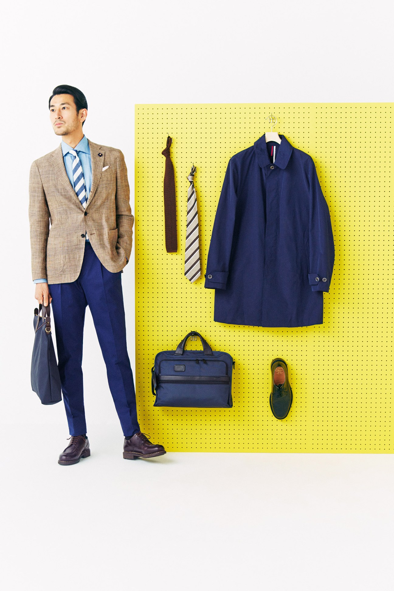 Man in beige Lardini jacket and navy trousers poses beside yellow pegboard displaying ties, navy coat, bag and shoes.