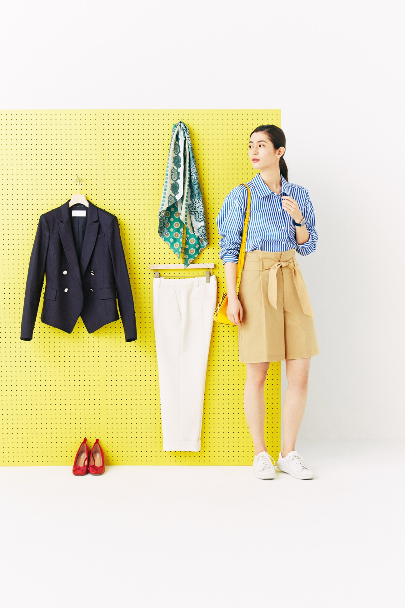 Woman styling spring wardrobe on yellow pegboard with navy blazer, striped shirt, and beige shorts.
