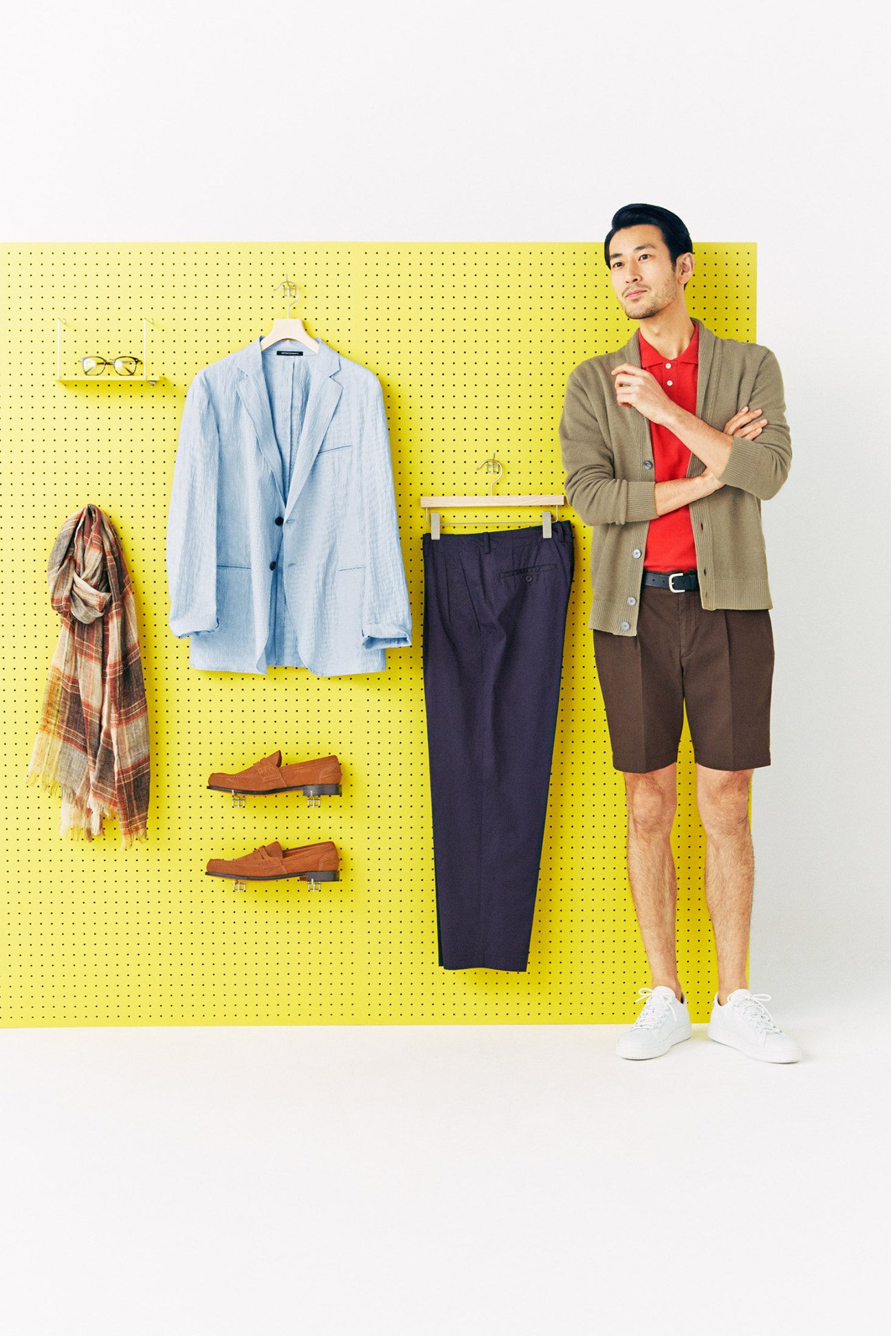 Man posing next to yellow pegboard displaying cardigan, shirt, shorts, trainers and accessories for spring wardrobe styling