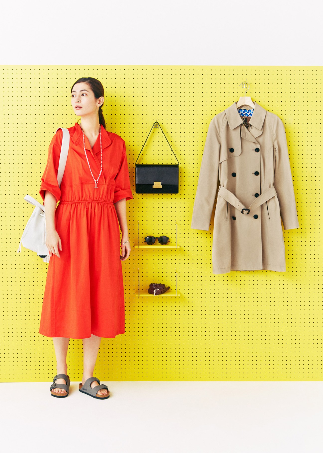 Woman in red dress and Birkenstock sandals poses next to clothing display board with trench coat and accessories against yellow pegboard.