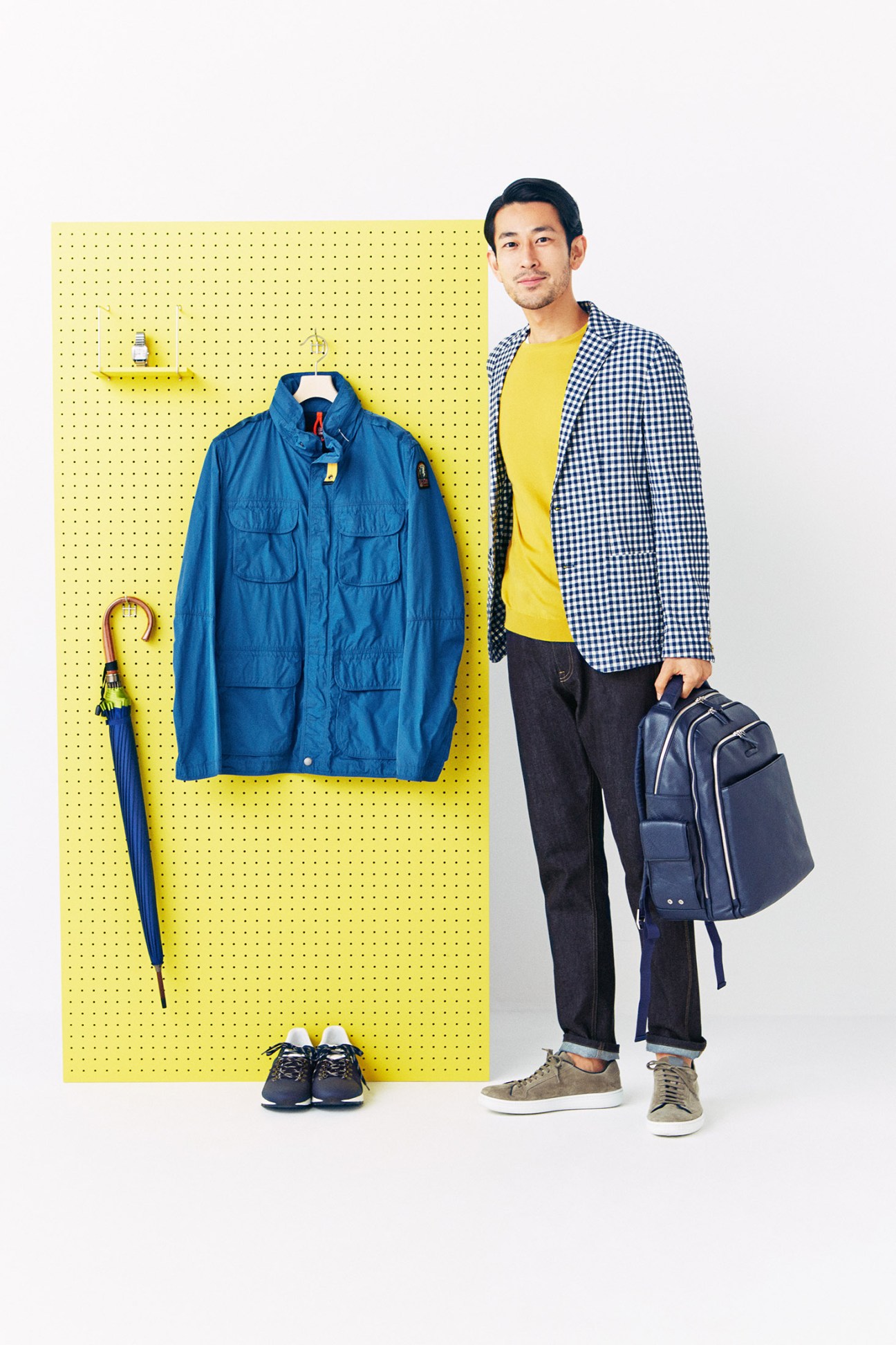 Male model in yellow pegboard setting wearing Brooksfield jacket, John Smedley jumper, Prada jeans and Church's trainers, holding Piquadro backpack.