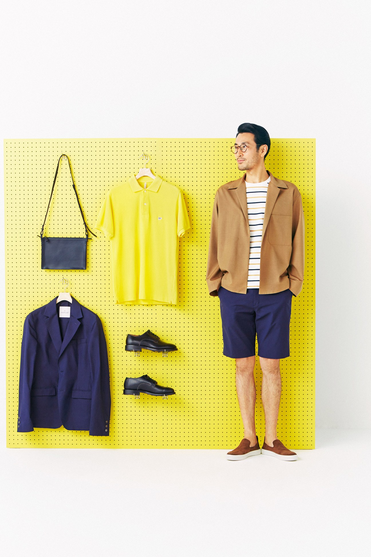Male model wearing camel jacket and navy shorts posed against yellow pegboard displaying coordinated spring wardrobe pieces.