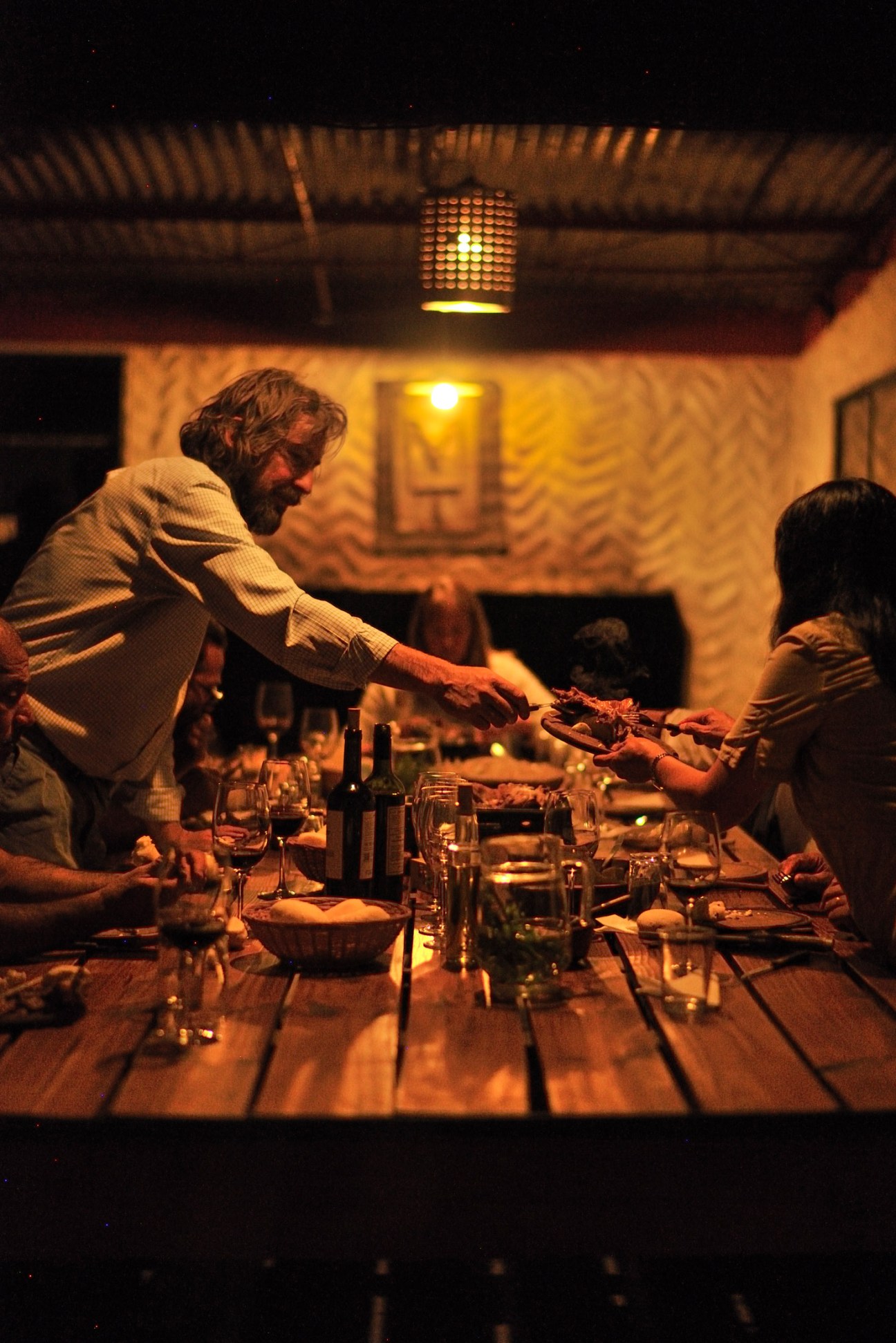 Man serving food to diners at a long candlelit table in a rustic stone room.