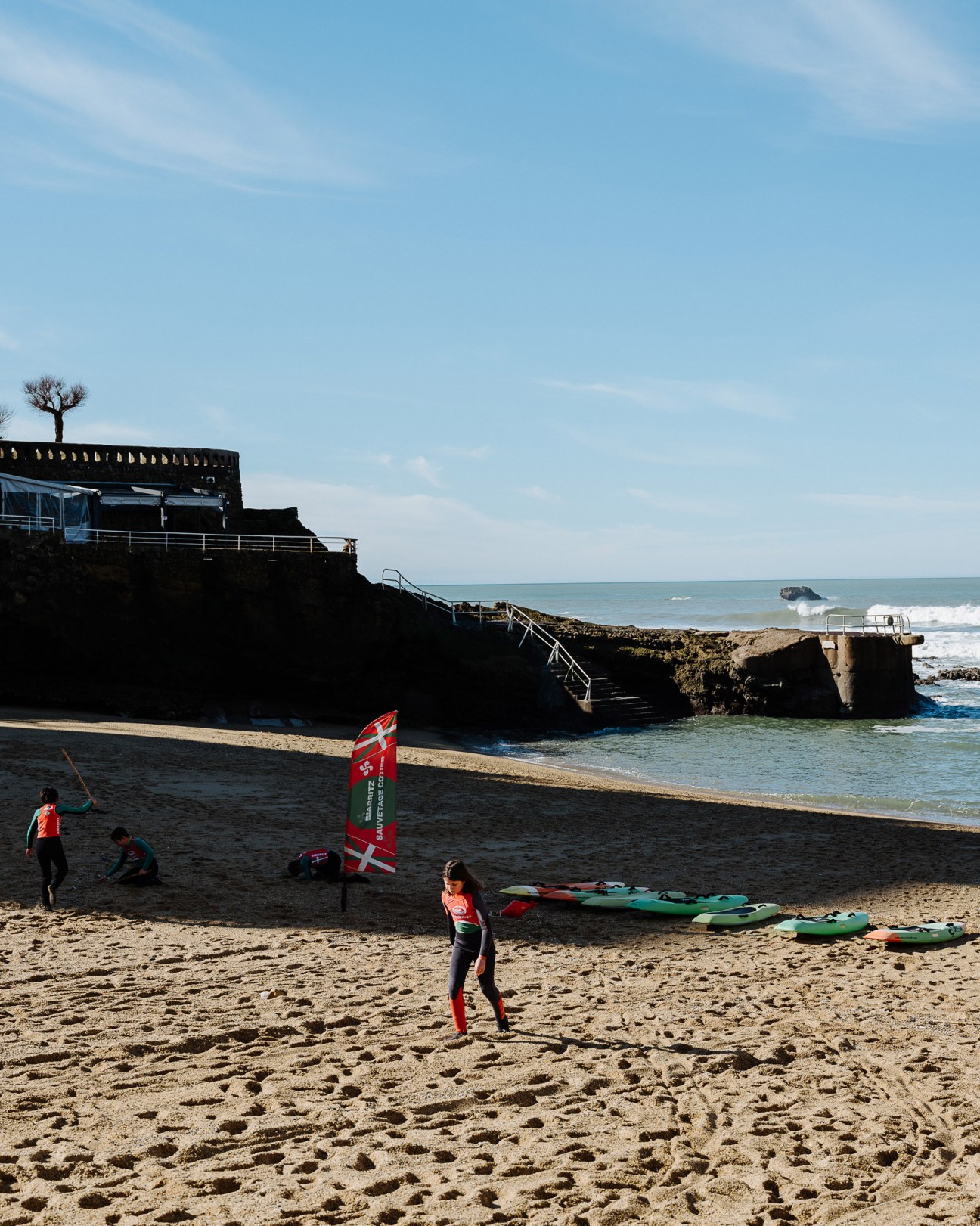 Kitesurfers on a sandy beach with black rocky cliffs and a building overlooking Plage du Port Vieux in Biarritz.