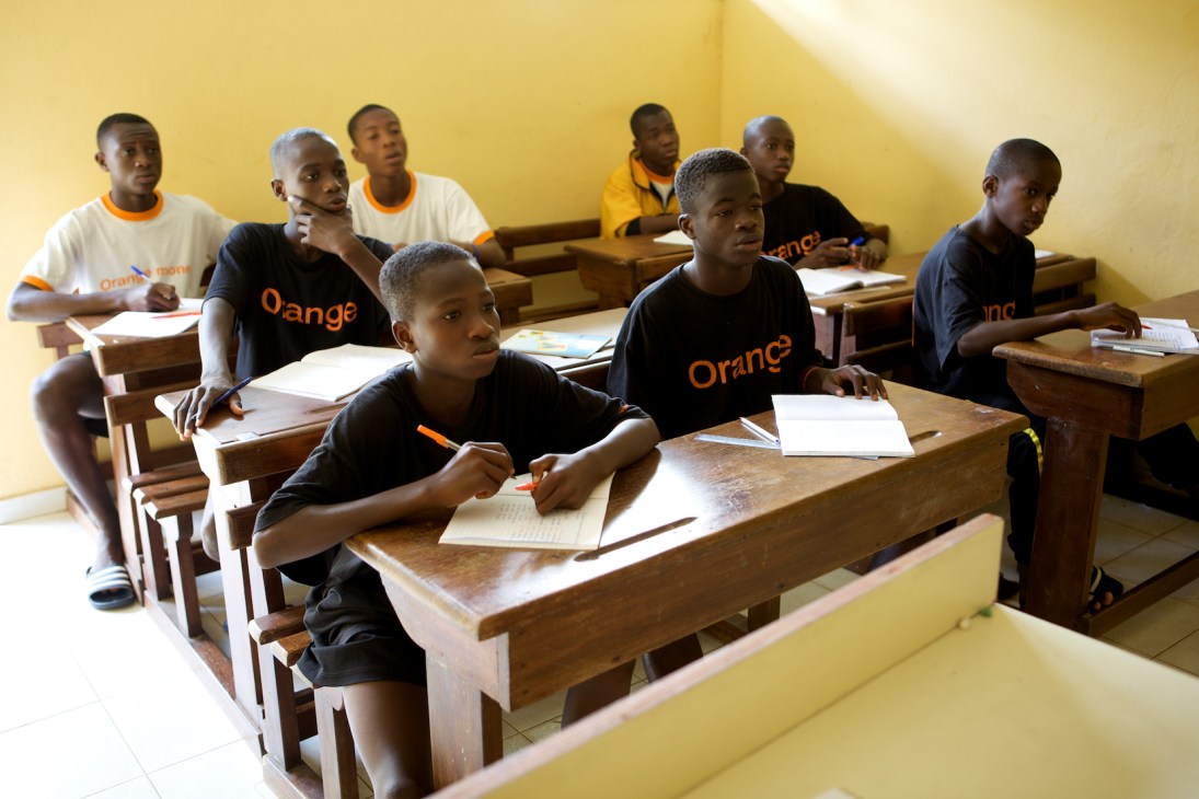 Young ASEC Mimosas academy students wearing Orange-branded shirts study at wooden desks in a yellow classroom.
