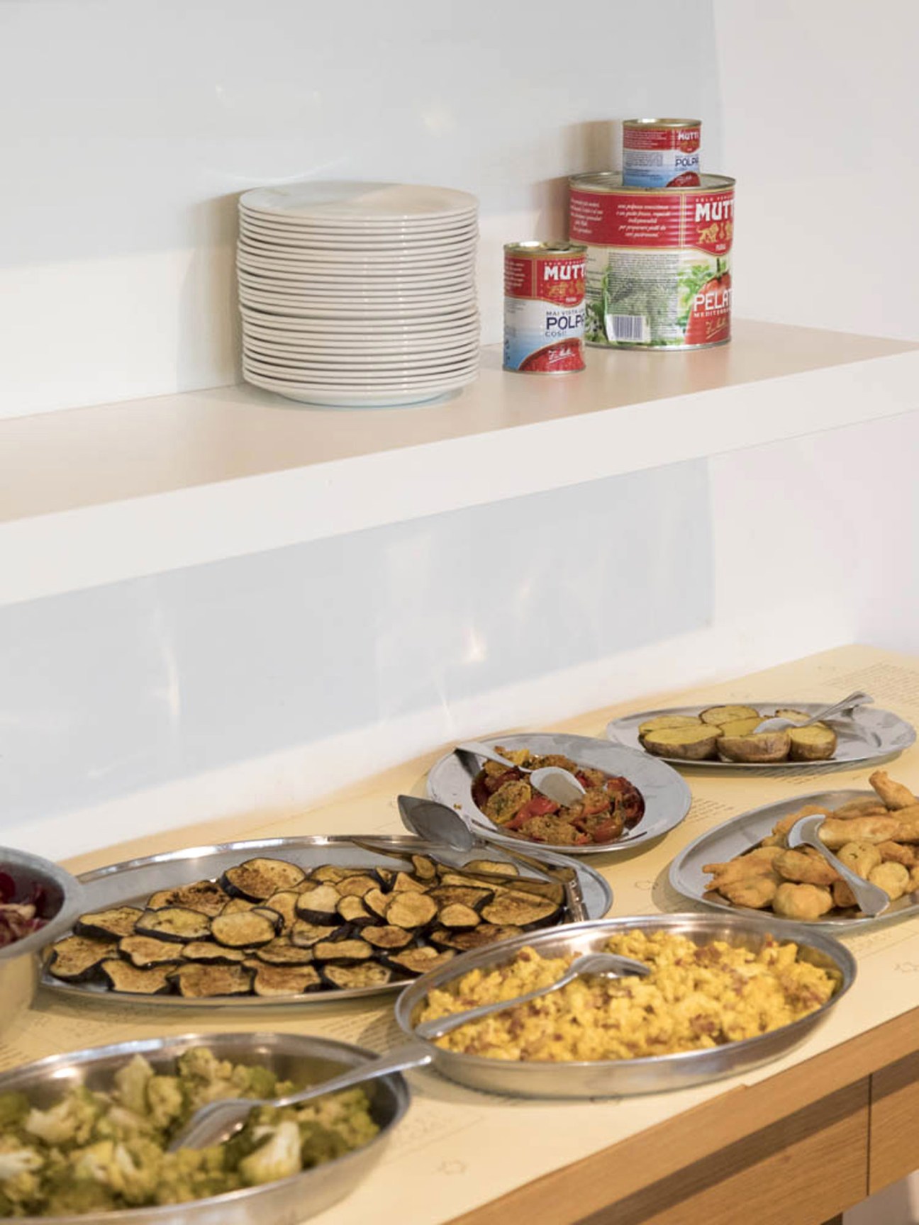 Fresh vegetables displayed on serving platters at a company buffet with canned goods on shelf above.