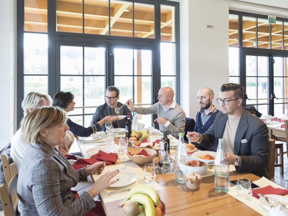 Business clients and staff dining together at a wooden table with large windows overlooking greenery at Brunello Cucinelli's company canteen.