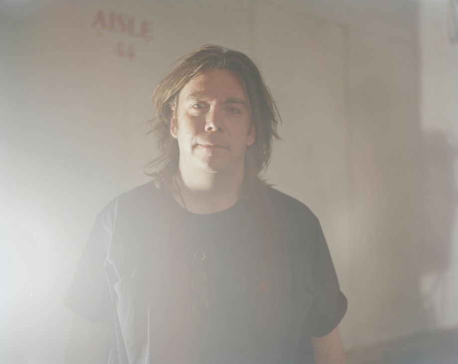 Dead of Night director Kevin Munroe, a man with shoulder-length hair wearing a dark t-shirt, photographed against a hazy light background.