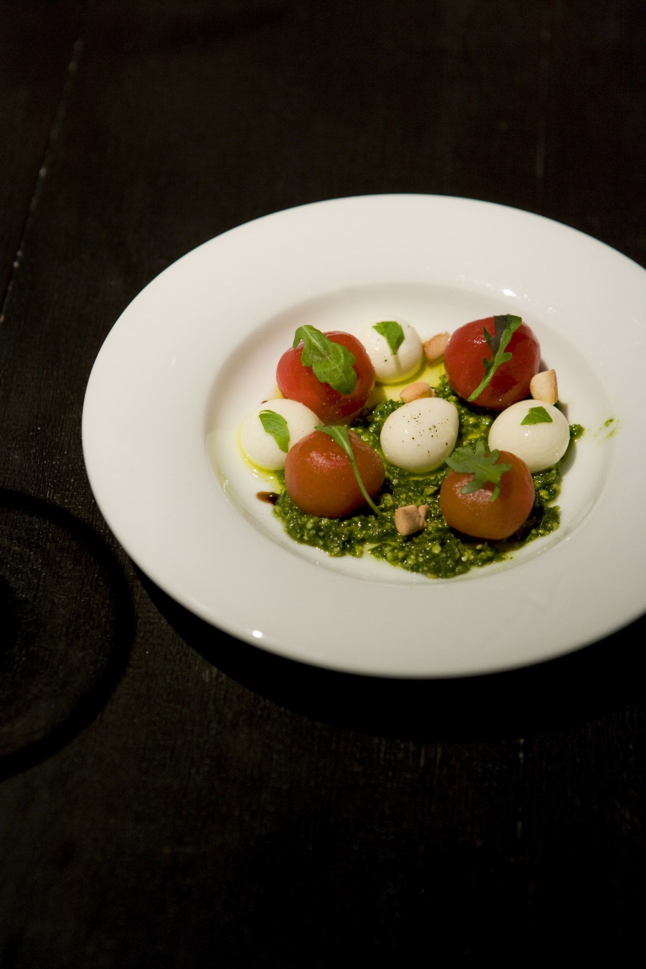 Plated caprese salad with tomato spheres, mozzarella balls, basil, and green sauce on white plate.