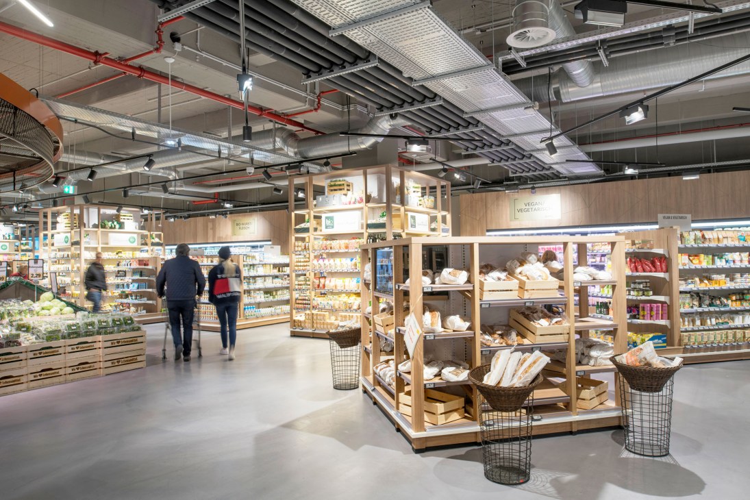 Bright, spacious interior of Zurheide supermarket with wooden shelving units, industrial ceiling, and shoppers browsing aisles.