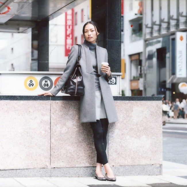 Woman in grey Stella McCartney coat and Drawer jumper standing against concrete wall in urban setting.