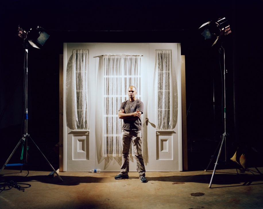 Co-producer Manu Gargi stands on a film set in front of a white door with arms crossed, flanked by professional lighting equipment.