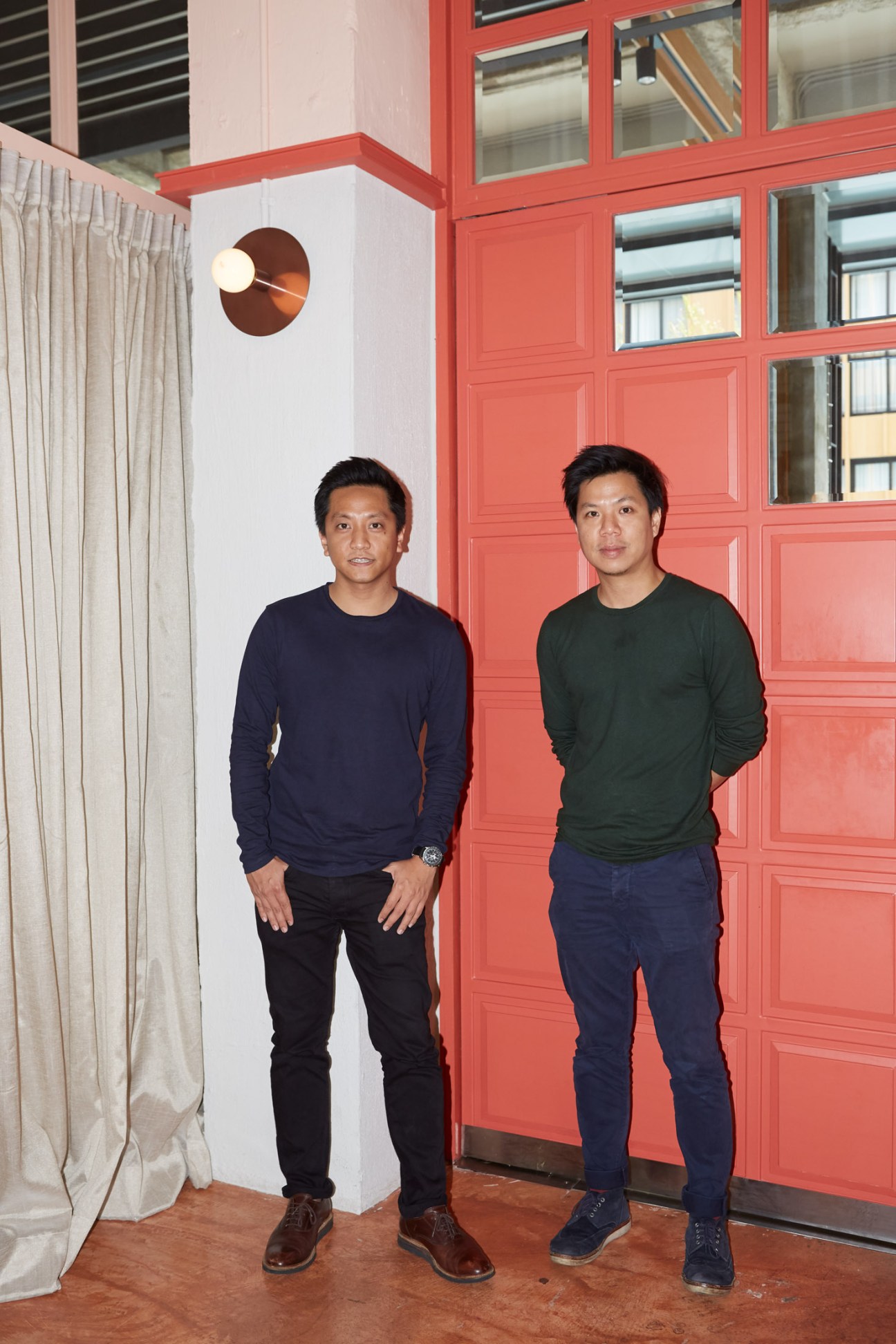Two men stand against a white wall with a red paneled door behind them in an interior space.