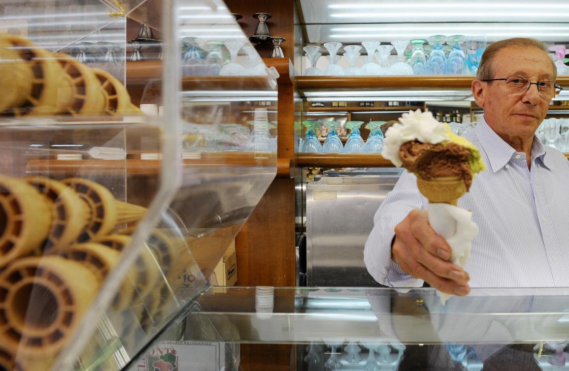 An older man with glasses in a light blue shirt holds a pastry inside a gelato shop display case.