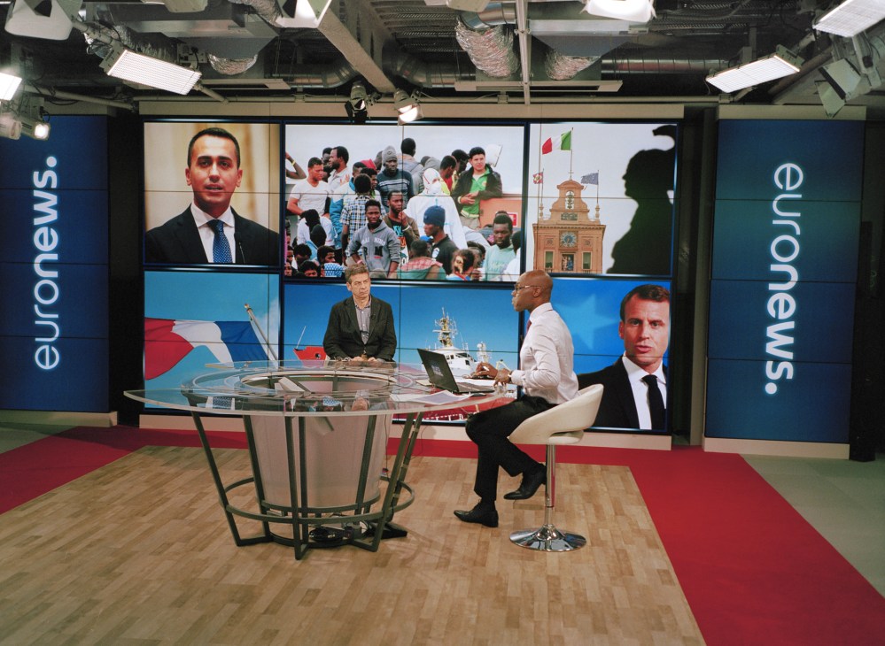Two presenters conduct an interview at a curved desk in a modern television studio with large screens displaying news content behind them.