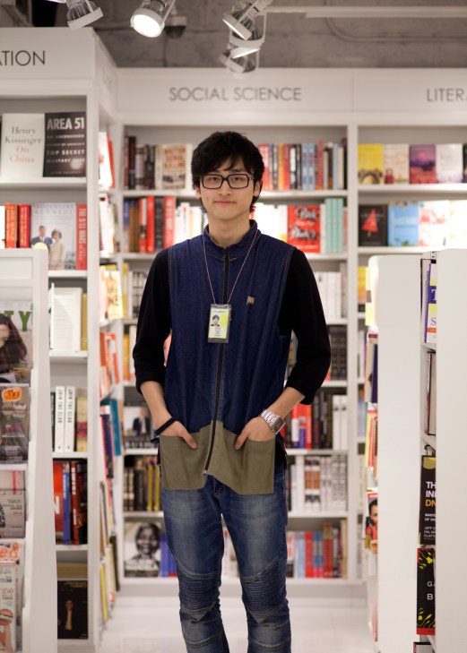 Young man wearing glasses and navy vest stands in front of well-stocked bookshelves at Page One airport kiosk.
