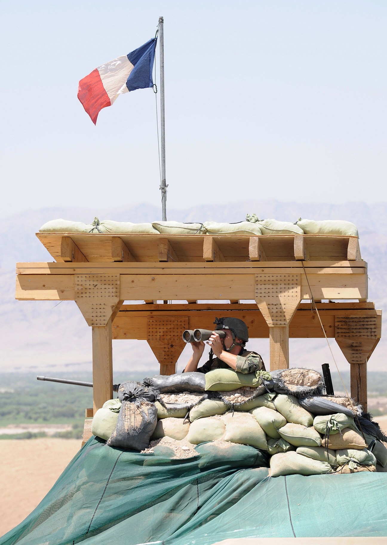 French soldier observing from fortified position with binoculars beneath French flag in Afghanistan.