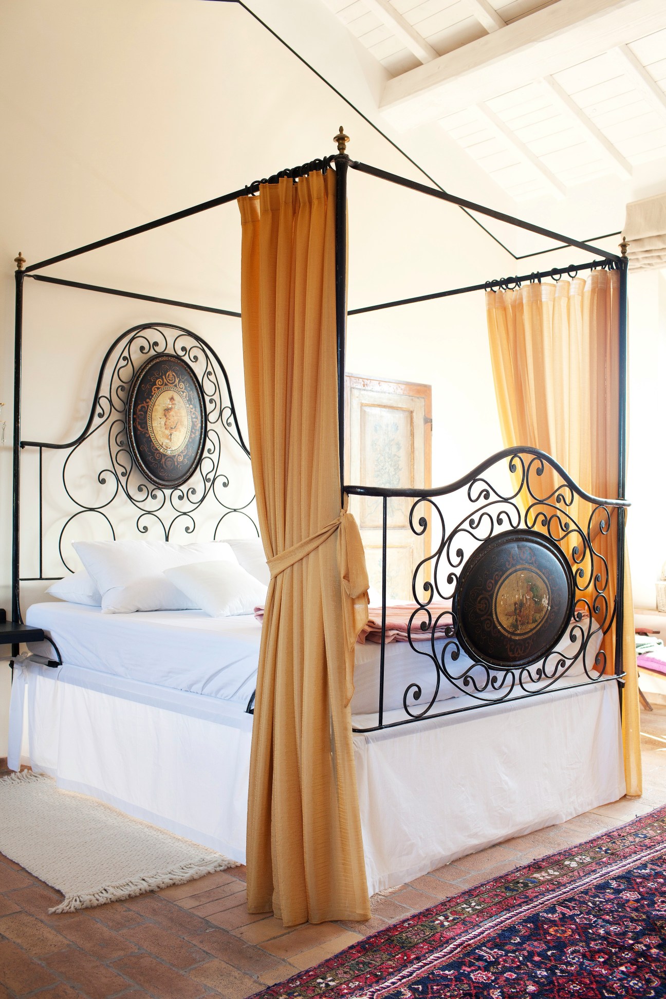 Medieval bedroom with ornate black iron four-poster bed, white linens, golden curtains and decorative wall medallion.