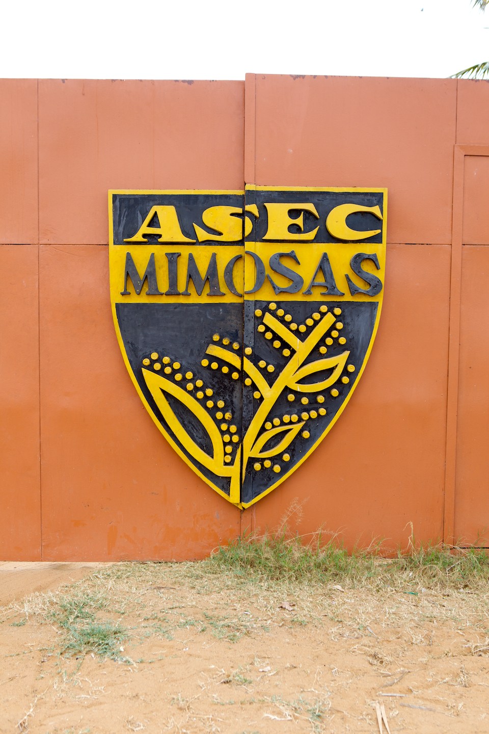 ASEC Mimosas football club badge with yellow and black shield design on terracotta wall.