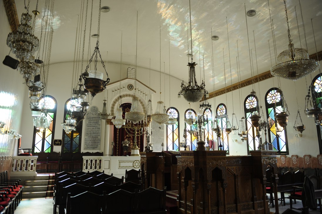 Interior of a synagogue with ornate hanging lamps, wooden pews, and stained glass windows.