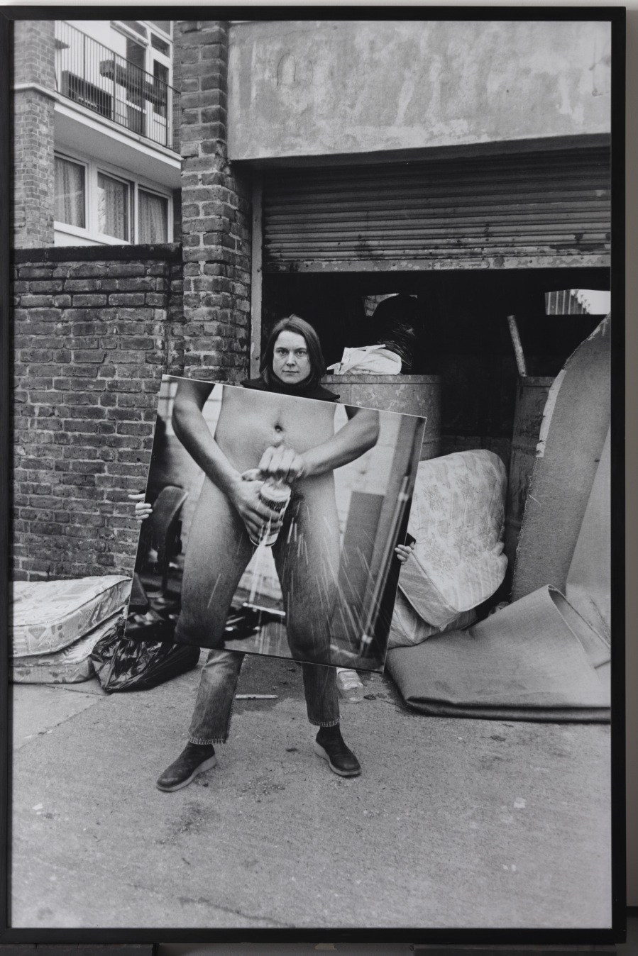 Black and white photograph of a person holding an explicit artwork outside a brick building.