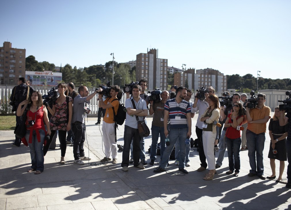 Large group of press photographers and journalists waiting outside Zaragoza Station with residential buildings in background.