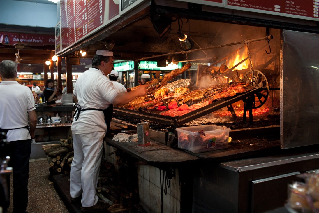 A chef in white uniform tends to meat roasting over an open flame at the port market.