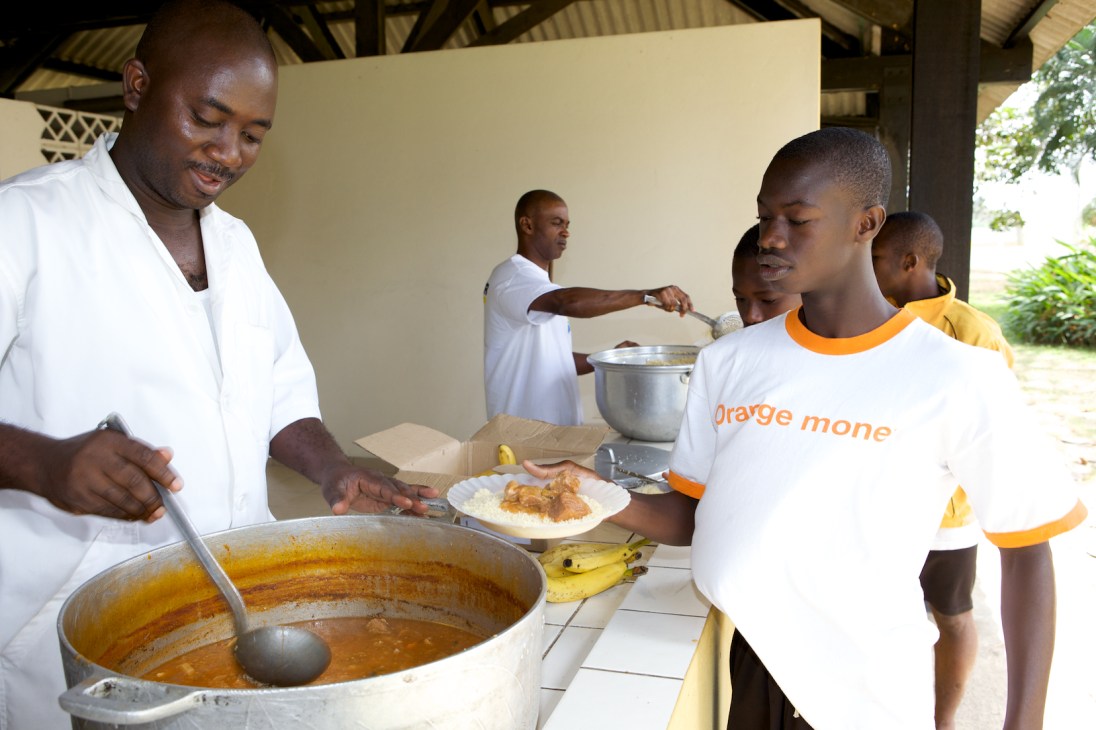 Young football academy students queue for dinner served by staff in a covered dining area.