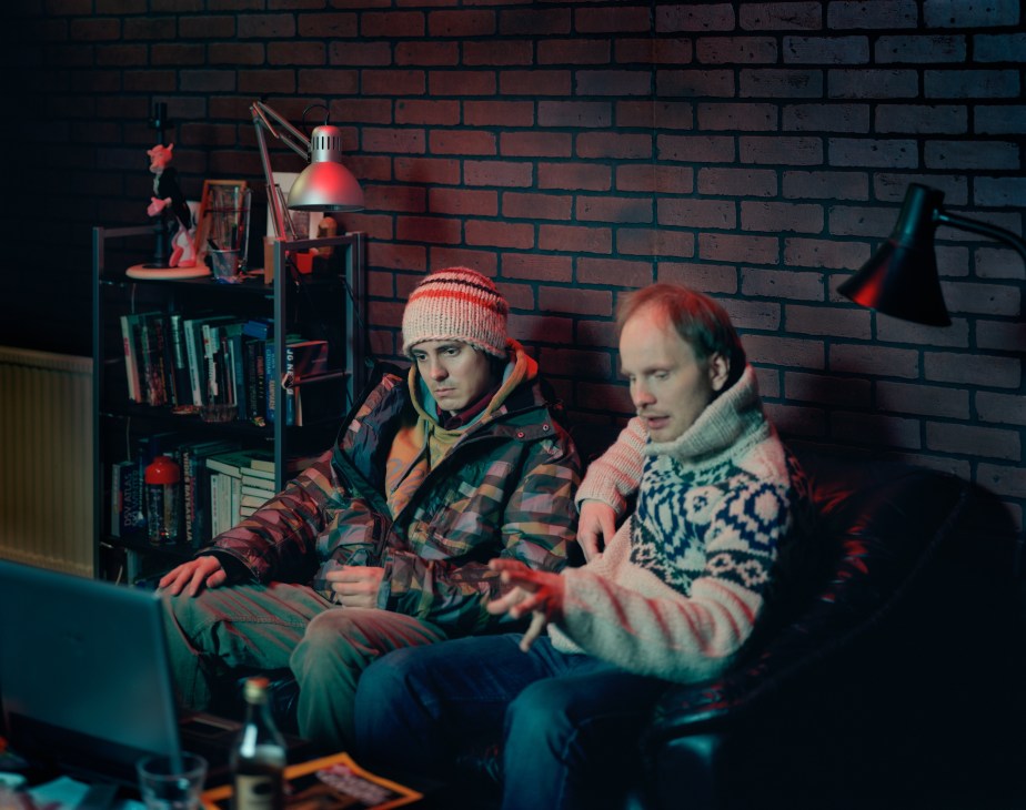 Two men discuss production in a studio workspace with brick walls, shelving, and red industrial lighting.