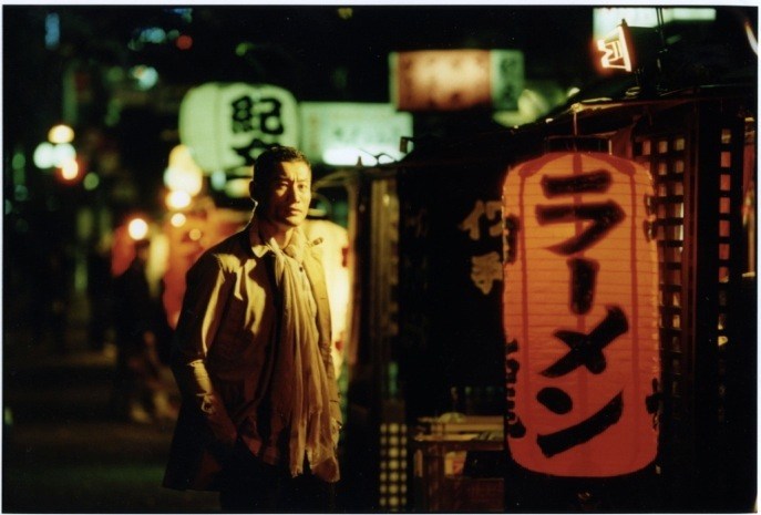 Model wearing Nanamica coat and Comme des Garçons trousers stands on a nighttime Fukuoka street lined with illuminated red lanterns.