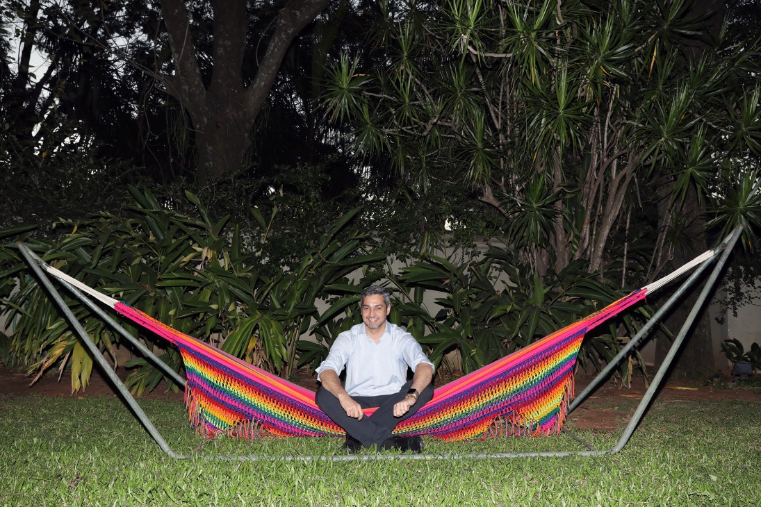 Man in light blue shirt sitting in colorful hammock in tropical garden setting.