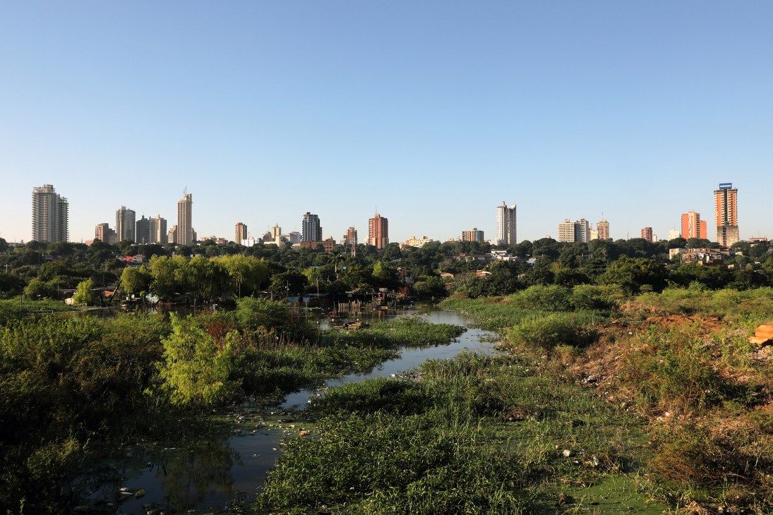 Asunción skyline with high-rise buildings rising above lush green vegetation and a winding waterway in the foreground.