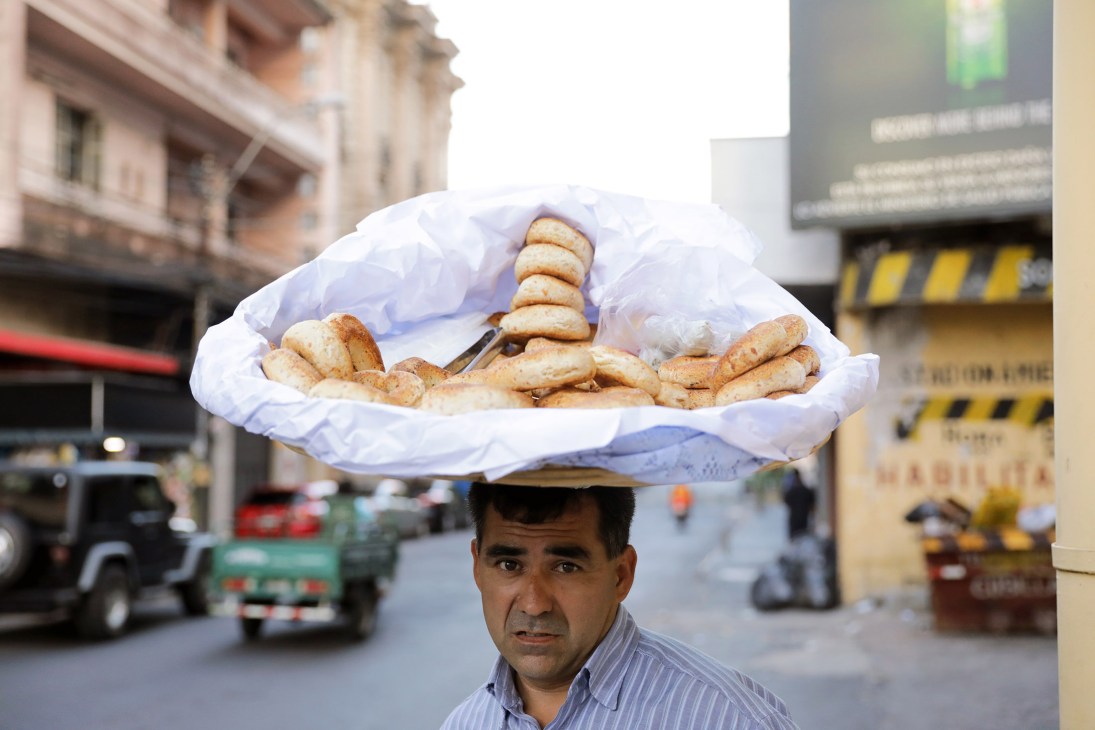 A vendor balances a large white paper cone filled with chipa bread on his head in a city street.