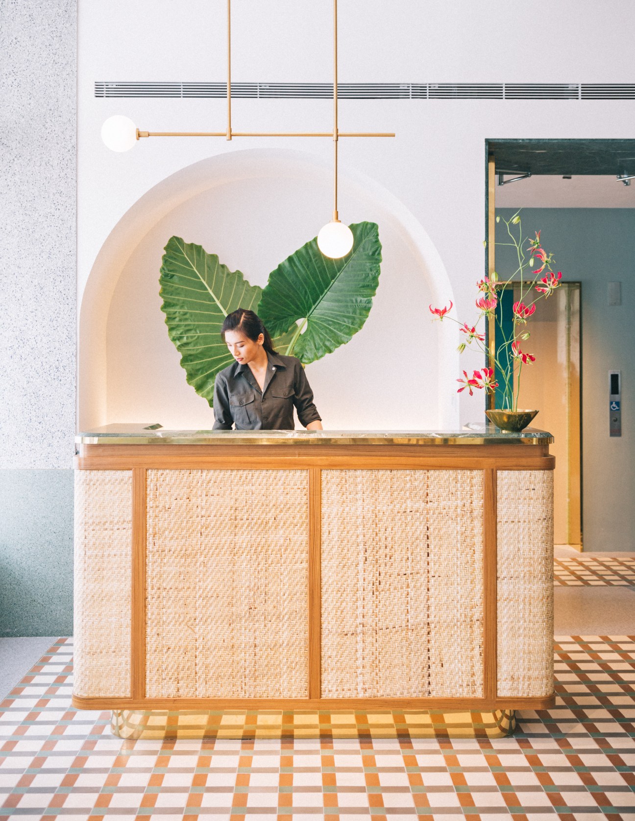A woman stands behind a woven wooden reception desk with large green tropical leaves and red flowers displayed behind her.