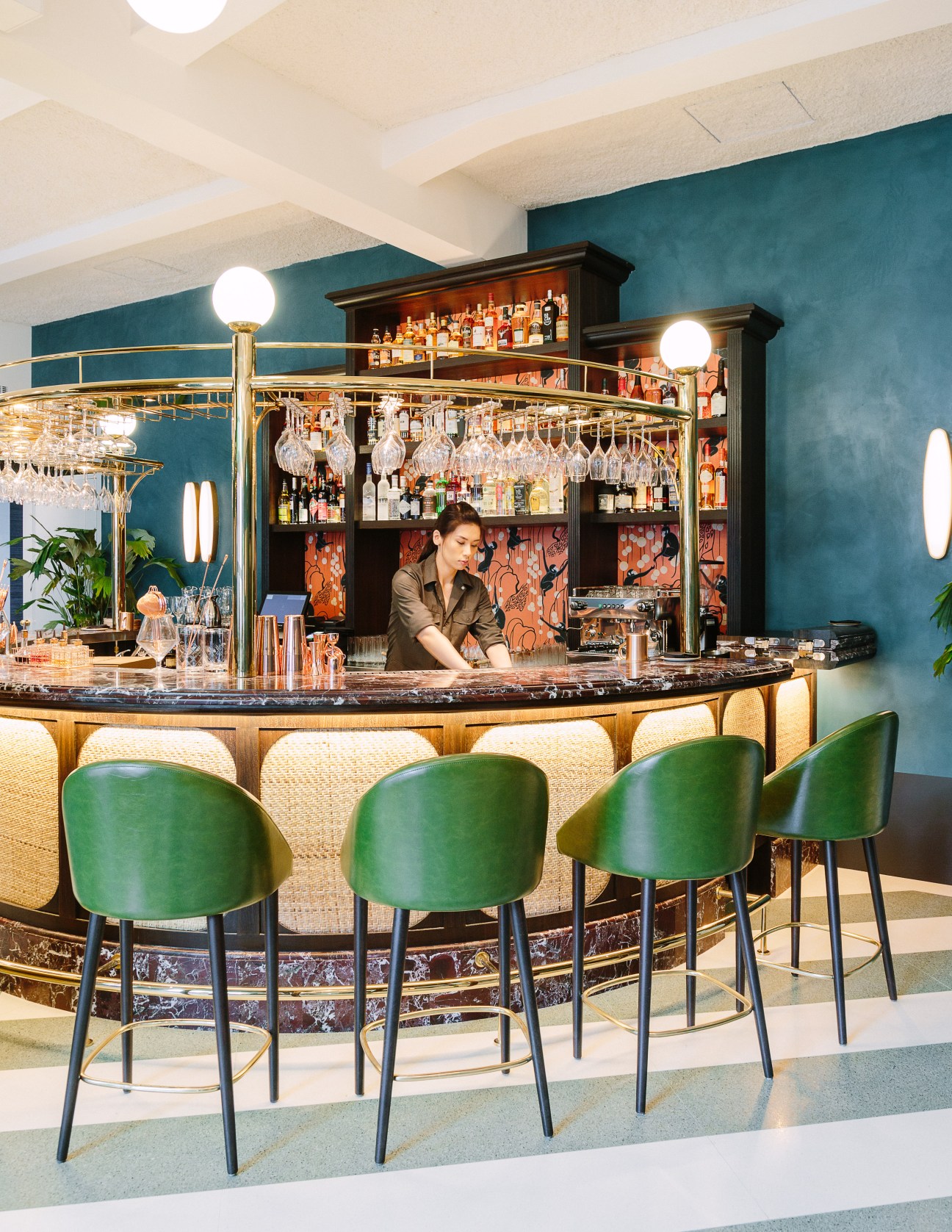 Bartender at well-stocked bar with teal walls, brass fixtures, and green bar stools at Straits Clan.