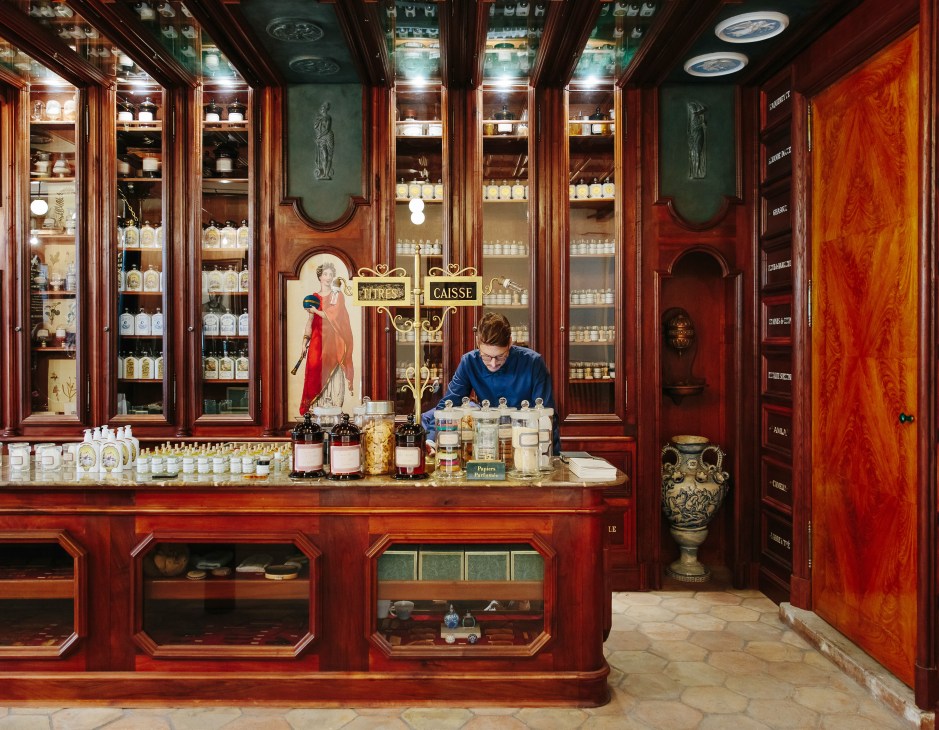 Staff member at wooden counter with glass-fronted cabinets displaying Buly 1803 fragrances and products