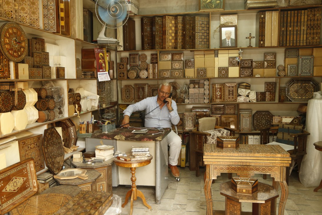 An older man on the phone in a workshop filled with ornate inlaid wooden boxes and traditional crafts.