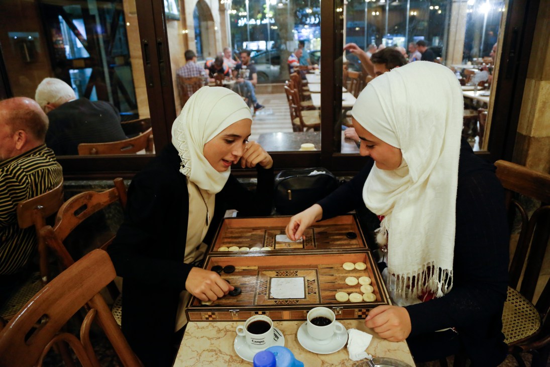 Two women in white hijabs playing backgammon and drinking Turkish coffee at a cafe table.